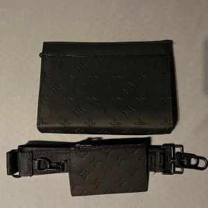 Svart axelväska med monogram - Snygg svart axelväska med präglat monogrammönster. Väskan har en justerbar axelrem och en liten avtagbar plånbok med dragkedja. Perfekt för att bära dina nödvändigheter med stil.