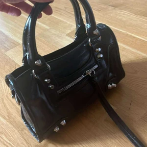 Svart handväska med nitar och dragkedja - Cool svart handväska i skinn med silvriga nitar och detaljer. Väskan har två handtag, en dragkedja framtill och dekorativa metalldetaljer. Perfekt för dig som gillar edgy stil och vill ha en väska som sticker ut.