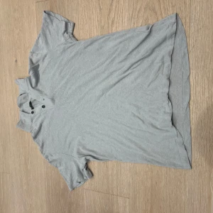 Ljusgrå piké från Massimo Dutti - Snygg ljusgrå pikétröja från Massimo Dutti i en mix av linne och bomull. Klassisk krage med två knappar framtill och korta ärmar. Perfekt för en clean och stilren look.