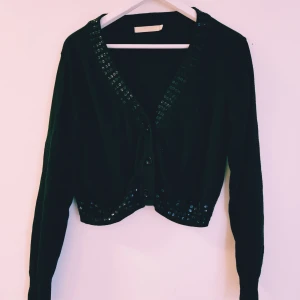 Svart kofta med paljettdetaljer från cappuccini strl XL - Svart kofta från Cappuccini Star i storlek XL. Croppad modell med långa ärmar och knappar framtill. Snygga svarta paljetter längs v-ringningen och nederkanten ger extra bling. Perfekt för dig som vill sticka ut lite extra.  Helt & fint skick dock tvättludd