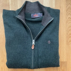 Mörkgrön kofta från Morris i merinoull - Snygg mörkgrön kofta från Morris i mjuk merinoull. Full dragkedja med läderdetalj, ribbade muddar och diskret logga broderad på bröstet. Perfekt till lager-på-lager och funkar året runt. Klassisk och stilren modell med normal passform.