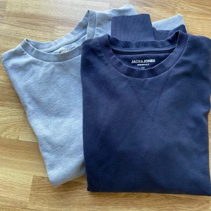 2 Jack & Jones crewneck tröjor XS - Två stilrena crewneck tröjor från Jack & Jones i storlek XS. En är mörkblå och den andra ljusgrå, båda med ribbade muddar och rund hals. Perfekta för en clean och enkel look. Tillverkade i mjuk bomull för skön känsla hela dagen.