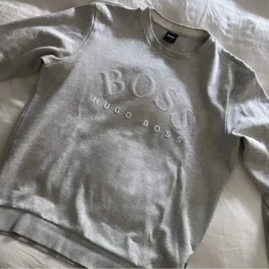 Grå sweatshirt från Hugo Boss Strl XL/L - Snygg grå sweatshirt från Hugo Boss med stort, upphöjt BOSS-tryck på bröstet. Tröjan har rund halsringning, långa ärmar och ribbade muddar. Perfekt för en clean och stilren look, funkar lika bra till jeans som till mjukisbyxor. I storlek XL men passar verkligen L också!