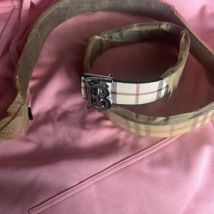 Burberry rutig bälte med silverdetalj - Snyggt bälte från Burberry med klassiskt rutigt mönster i beige. Bältet har en stilren silverfärgad spänne med Burberrys ikoniska logga. Perfekt accessoar för att lyfta din outfit med en touch av lyx.                      Helt nytt endast privat