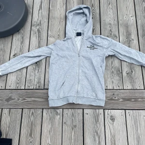 Ljusgrå hoodie från Peak Performance - Ljusgrå hoodie från Peak Performance med dragkedja och huva. Tröjan har två fickor framtill och diskret logga på bröstet. Tillverkad i mjuk bomullsblandning, perfekt för chill dagar eller när du vill ha en enkel och clean look.