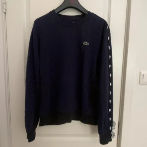 Mörkblå sweatshirt från Lacoste - Mörkblå sweatshirt från Lacoste med klassisk krokodillogga på bröstet och vita Lacoste-loggor längs ärmarna. Tröjan har rund hals, långa ärmar och ribbade muddar. Perfekt för dig som gillar stilren streetwear med sportig vibe.