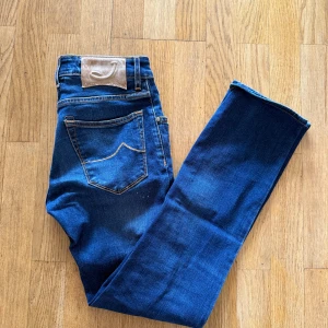 Jacob Cohën Nick blå jeans - Snygga blå jeans från Jacob Cohën, modell Nick. Klassisk femficksdesign med orange kontrastsömmar, knäppning med knappar och diskret logga på bakfickan. Jeansen har en rak passform och är tillverkade i mjukt bomullsjeansmaterial.