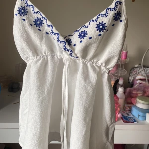 Vit topp - Supersöt vit linnetopp från SHEIN med blå broderade blommor längs v-ringningen. Tunt, lätt material med smala axelband och markerad midja med knyt. Perfekt för varma dagar och ger en fräsch sommarkänsla.