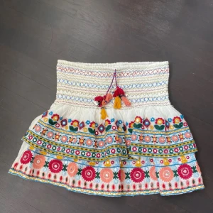 Broderad kortkjol med volang och tofsar - Färgglad kortkjol i bomull med broderade blommor i rött, gult, blått och grönt. Kjolen har volang, smock i midjan och dekorativa tofsar framtill. Perfekt för dig som gillar boho-stil och detaljer.