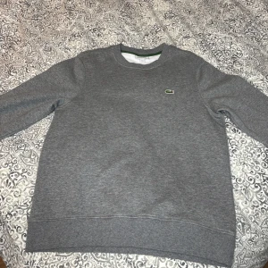 Grå sweatshirt från Lacoste - Klassisk grå sweatshirt från Lacoste med rund halsringning och den ikoniska gröna krokodilloggan broderad på bröstet. Tröjan har ribbade muddar vid ärmslut och nederkant samt en relaxed passform. Perfekt för en chill och stilren look. Helt ny oanvänd!