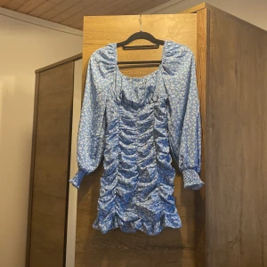 Blommig blå kortklänning från SHEIN - Supersöt blå kortklänning från SHEIN med vita blommor, långa puffärmar och smock vid ärmslut. Klänningen har rynkade detaljer framtill, volangkant nertill och dragkedja i sidan. Perfekt för dig som gillar romantisk och trendig stil.