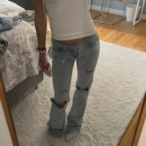 Ljusblå bootcut jeans med egengjorda slitningar - Säljer ett par ljusblå bootcut jeans med slitna detaljer över knäna och cool broderad röd detalj på bakfickorna. Låg midja och klassisk femficksmodell. Perfekta för dig som gillar en avslappnad och trendig look. Storlek 32.