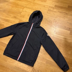 Moncler vindjacka - Säljer en svart vindjacka från Moncler  och snedställd dragkedja i rött, vitt och blått. Jackan har Moncler-logga på ärmen och två fickor framtill. Perfekt för dig som vill ha en stilren och sportig look. Jackan är helt ny den har använts en gång bara, priset kan diskuteras. Köparen står för frakten, storleken är S men passar Även M.