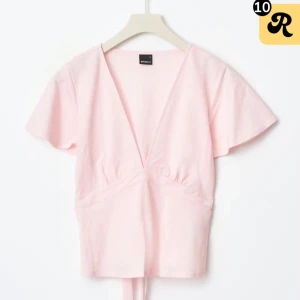 Ljusrosa omlottopp med knytband - Supersöt ljusrosa omlottopp från Gina  med v-ringning och korta ärmar. Toppen har ett knytband i midjan som ger en fin siluett. Perfekt att styla med jeans eller kjol för en fräsch look.