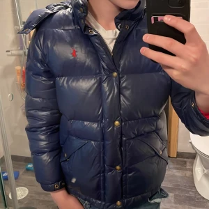 Blå dunjacka från Polo Ralph Lauren - Säljer en blå dunjacka från Polo Ralph Lauren med huva och dragkedja samt knappar. Jackan har två fickor framtill och den klassiska röda loggan på bröstet. Perfekt för kyliga dagar. Det är storleken 14-16 år men passa nån runt 160-170har två väldigt små hål på en av armarna men inget man tänker på nå mycket. Hör av er om ni har frågor eller funderingar🤩