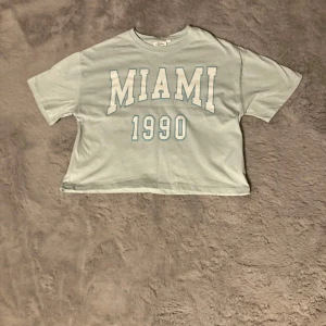 Ljusblå croppad t-shirt med MIAMI-tryck - Säljer en ljusblå croppad t-shirt med stort vitt MIAMI 1990-tryck på framsidan. T-shirten har korta ärmar och en avslappnad passform, perfekt för en cool och sportig stil.