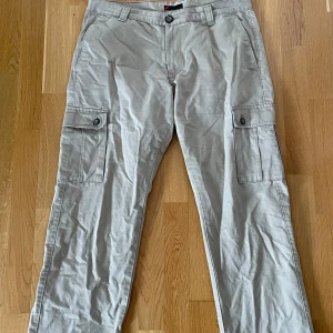 Beiga cargobyxor från Dressmann - Säljer ett par beiga cargobyxor från Dressmann med klassiska sidofickor med lock och knapp. Byxorna har normal passform och är tillverkade i bomull, perfekta för en avslappnad stil.