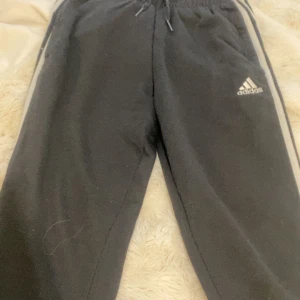 Svarta träningsbyxor från Adidas med vita ränder - Svarta Adidas träningsbyxor med klassiska vita ränder längs sidorna och dragsko i midjan. Byxorna har Adidas-logga på vänster ben och är perfekta för sportiga tillfällen. Bekväm passform och elastisk midja.
