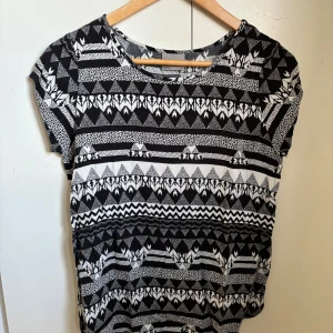 Svartvit mönstrad topp från Vero Moda - Säljer en svartvit topp från Vero Moda med grafiskt mönster i olika former och ränder. Toppen har rund halsringning och är kortärmad. Perfekt för dig som gillar unika mönster och en avslappnad stil.