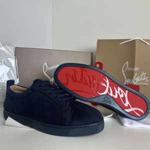 Christian Louboutin Skor - Säljer ett par svarta sneakers från Christian Louboutin med klassisk röd sula och diskret broderad logga på hälen. Skorna har snörning och är tillverkade i mocka med en platt sula. Perfekta för dig som gillar stilrena och exklusiva sneakers.