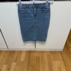 Blå jeanskjol med rå kant - Säljer en klassisk blå jeanskjol med rå, fransig nederkant och femficksmodell. Kjolen har normal passform och går till knäna. Perfekt till vardags med en t-shirt eller stickad tröja.
