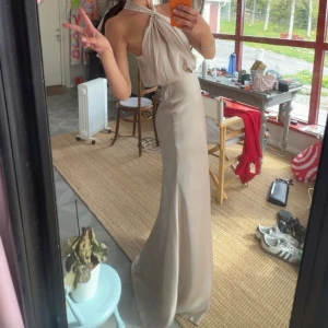 Champagnefärgad långklänning med halterneck - Säljer en elegant långklänning i champagnefärg med halterneck och draperad överdel. Klänningen har en figurnära passform och är tillverkad i ett glansigt, mjukt material som ger en lyxig känsla. Perfekt för bal eller fest!