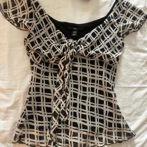 Svart och vit mönstrad blus från H&M - Säljer en svart och vit blus från H&M med rutigt mönster och knyt framtill. Blusen har korta ärmar och lätt v-ringning. Tunt och luftigt material