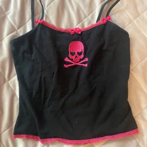 Svart linne med dödskalle och rosa spets - Säljer ett svart linne med smala axelband, dekorerat med rosa spets och små rosetter. Framtill finns ett broderat dödskallemotiv i rosa. Perfekt för dig som gillar alternativ stil!