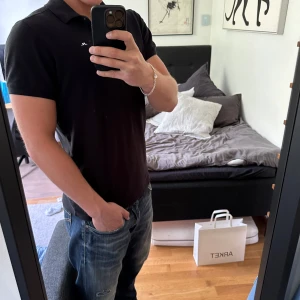 Svart piké t-shirt från J.Lindeberg - Säljer en stilren svart piké t-shirt från J.Lindeberg med klassisk krage och korta ärmar. Perfekt för en clean och enkel look. Liten logga på bröstet. Passar dig som gillar en normal passform.