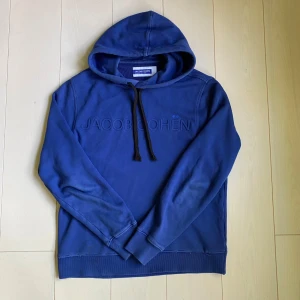Blå hoodie från Jacob Cohën - Säljer en blå hoodie från Jacob Cohën med broderad logga på bröstet och svarta snören i huvan. Tröjan har långa ärmar och ribbade muddar vid ärmslut och nederkant. Perfekt för en avslappnad stil.