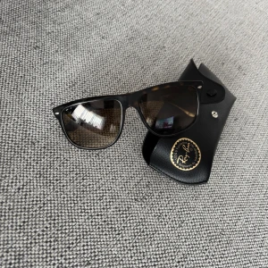 Ray-Ban Wayfarer solglasögon - Snygga Ray-Ban Wayfarer solglasögon med klassisk svart och brunmelerad båge. Glasögonen har en tidlös design och kommer med originalfodral. Perfekt accessoar för soliga dagar.