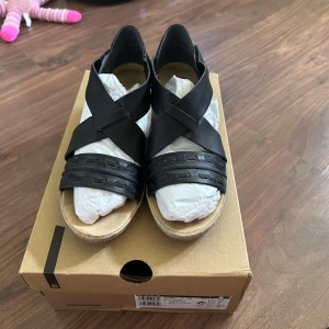 Svarta sandaler med kilklack från Rieker - Snygga svarta sandaler från Rieker med korsade remmar i syntet och öppen tå. Skorna har en bekväm kilklack och platt sula framtill. Perfekta för varma dagar och ger en stilren look till din outfit. Endast använda 1 gång. 