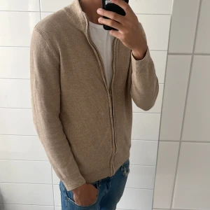 Beige fullzip - Säljer en beige fullzip från Indicode i storlek Large. Tröjan har dragkedja framtill och hög krage, perfekt för lager-på-lager. Enkel och stilren design som passar till det mesta.