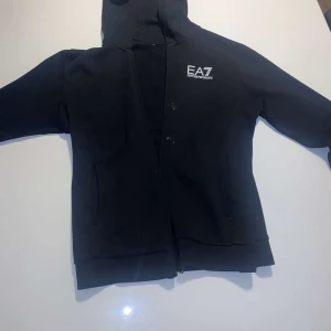 Svart hoodie från Emporio Armani EA7 - Svart hoodie med dragkedja från Emporio Armani EA7. Klassisk design med huva och logga på bröstet. Perfekt för en avslappnad och stilren look.