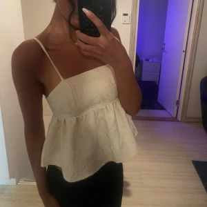 Vit spetslinne med volang - Säljer en söt vit linnetopp med tunna axelband och volang nertill. Toppen har ett vackert spetsmönster och är perfekt för varma dagar. Passformen är luftig och modellen är croppad.