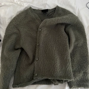 Olivgrön kofta med knappar - Mysig olivgrön kofta med v-ringning och stora knappar framtill. Tröjan har lång ärm och en lite lurvig, stickad struktur som ger en avslappnad look. Perfekt att slänga på sig över en t-shirt eller topp.