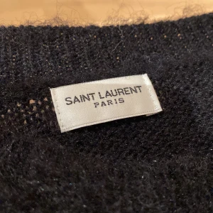 Svart fluffig tröja från Saint Laurent - Säljer en svart, fluffig stickad tröja från Saint Laurent med vit text framtill. Tröjan har lång ärm och en oversized passform, perfekt för dig som vill ha något både snyggt och bekvämt. Ikonisk logga framtill. 