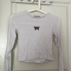Vit långärmad croppad tröja med fjäril från Pull&Bear - Vit långärmad croppad tröja från Pull&Bear med ett litet tryck av en lila och svart fjäril på bröstet. Enkel och stilren design som passar perfekt till jeans eller kjol.. storlek S