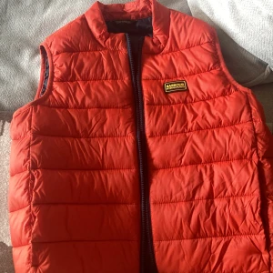 Röd dunväst från Barbour International - Säljer en röd dunväst från Barbour International med quiltad design och dragkedja framtill. Västen har en klassisk passform och Barbour-logga på bröstet. Perfekt att slänga över en hoodie eller tröja för extra värme.