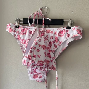 Vit och rosa blommig bikini - Snygg bikini i vitt med rosa blommönster. Toppen har knytbara band och är vadderad. Självklart oanvända, bara legat undanstoppade i en garderob.