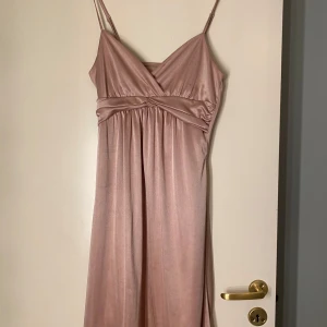 Champagnefärgad långklänning med smala axelband - Säljer min sidenrosa klänning med smala axelband och v-ringad omlott framtill. Klänningen har ett fint fall och är tillverkad i siden som ger en lyxig känsla. Köpt från veromoda ordnarie pris 499kr