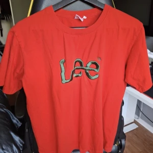 Röd t-shirt från Levi's med broderad Lee-logga - Säljer en röd t-shirt från Levi's med broderad Lee-logga i grönt och svart på bröstet. T-shirten har rund halsringning och korta ärmar. Perfekt för en avslappnad stil. Tröjan passar som M.