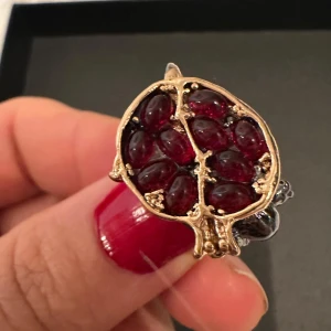 Ring i guldton med röda stenar - Unik ring i guldtonad metall med ett dekorativt motiv som liknar en granatfrukt fylld med djupröda, ovala stenar. Ringen har en konstnärlig och iögonfallande design som verkligen sticker ut.