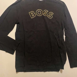 Svart långärmad tröja från BOSS - Säljer en svart långärmad tröja från BOSS med gul text på bröstet och liten logga i nacken. Tröjan har en klassisk passform och är tillverkad i mjuk bomull. Perfekt för en stilren och enkel look.