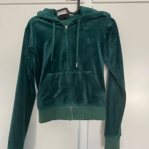 Grön velour hoodie från Juicy Couture - Mörkgrön hoodie i mjuk velour från Juicy Couture med dragkedja och huva. Klassisk modell med fickor framtill och ribbade muddar. Perfekt för en avslappnad och trendig look. Den har gått sönder vid dragkedjan
