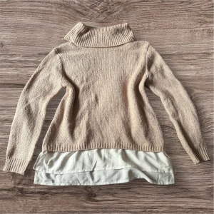Beige stickad polotröja med vit skjortkant från Love Knitwear - Säljer en beige stickad polotröja från Love Knitwear med lång ärm och hög krage. Tröjan har en snygg vit skjortkant nedtill som ger en lager-på-lager-look. Perfekt för en stilren och trendig outfit.