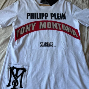 Vit Philipp Plein Scarface t-shirt - Säljer en vit t-shirt från Philipp Plein med stora tryck och detaljer inspirerade av Scarface och Tony Montana. T-shirten har korta ärmar, svart logga på ryggen och glittriga stenar på texten framtill. Perfekt statement-plagg för dig som gillar unika designer.