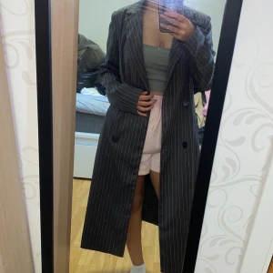 Grå långrandig kappa - Säljer en lång, grå kappa med vita smala ränder. Klassisk passform med långa ärmar och knappar framtill. Perfekt för dig som gillar en stilren och tidlös look.