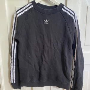 Svart sweatshirt från Adidas - Klassisk svart sweatshirt från Adidas med vita ränder längs ärmarna och liten Adidas-logga på bröstet. Köpt i USA för några år sen men knappt använd.
