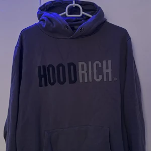 Mörkgrå hoodie från Hoodrich - Säljer en mörkgrå hoodie från Hoodrich med stor logga i svart och grått framtill. Tröjan har huva med dragsko och en stor magficka. Perfekt för dig som gillar streetwear och vill ha en bekväm och snygg tröja.